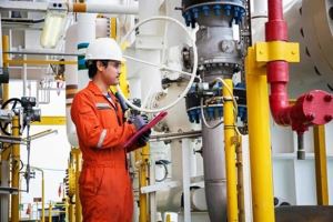 A safety worker wearing orange coveralls and a hard hat in an oil and gas facility reviews a checklist on a clipboard.